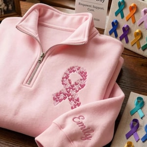 May include: A light pink pullover sweatshirt with a quarter-zip neckline and embroidered pink ribbon design. The sleeve is embroidered with the name "Emily". The image also shows a display of colorful ribbons and a card that reads "Empower. Support. Cure. 2026."