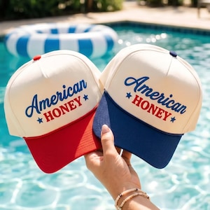 May include: Two baseball caps with cream-colored crowns and structured panels. One cap features a red brim and red text reading American HONEY with blue stars, while the other has a navy blue brim and blue text reading American HONEY with red stars.
