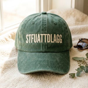 May include: Green baseball cap with the text "STFUATTDLAGG" in white block letters. The hat is made of a soft, worn fabric and has a curved brim. Sunglasses and greenery are visible in the background.