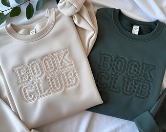 May include: Two crewneck sweatshirts, one beige and one dark green, are displayed. The beige sweatshirt has the words "BOOK CLUB" embroidered in a collegiate style. The dark green sweatshirt also has "BOOK CLUB" embroidered on it. The sweatshirts are on a white surface.