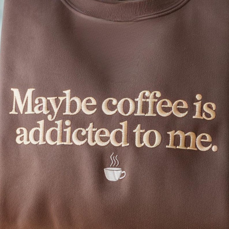May include: Brown sweatshirt with the embroidered phrase "Maybe coffee is addicted to me." The text is in a light beige color. A small white coffee cup with steam is embroidered below the text. The sweatshirt is a solid color.