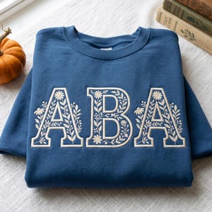 May include: A blue sweatshirt with the embroidered letters "ABA" in a floral design. The letters are outlined in white and filled with delicate floral and leaf patterns. A small pumpkin and books are in the background.