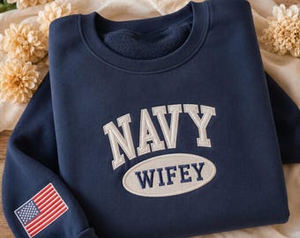 May include: Navy blue sweatshirt with the word "NAVY" embroidered in white block letters above "WIFEY" in an oval. An American flag patch is on the left sleeve. The sweatshirt is folded on a cream-colored surface.