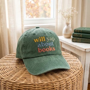 May include: A green baseball cap with the embroidered text "will yap about books" in yellow, blue, and red. The hat is sitting on a woven wicker stool. A window and books are in the background.