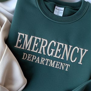 Op de afbeelding: Een teal groene sweatshirt met de woorden "EMERGENCY DEPARTMENT" geborduurd in cr&egrave;mekleurig garen. De sweatshirt heeft een ronde hals en een label aan de binnenkraag. Het kledingstuk is op een lichtbeige oppervlak gevouwen.