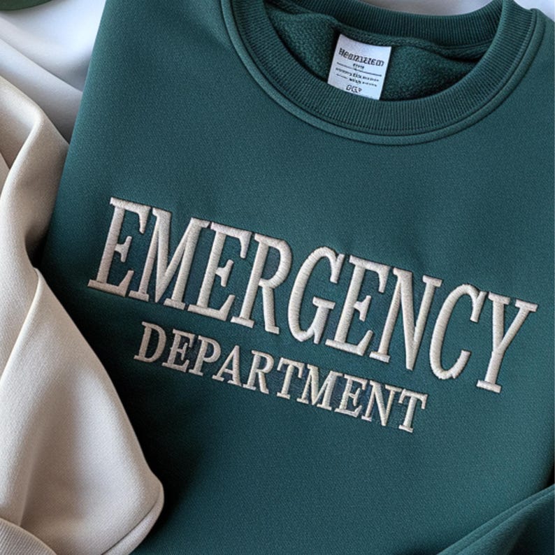 Op de afbeelding: Een teal groene sweatshirt met de woorden "EMERGENCY DEPARTMENT" geborduurd in cr&egrave;mekleurig garen. De sweatshirt heeft een ronde hals en een label aan de binnenkraag. Het kledingstuk is op een lichtbeige oppervlak gevouwen.