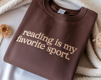 May include: A brown sweatshirt with the embroidered text "reading is my favorite sport." The crew neck sweatshirt is folded and resting on a white, textured surface. A dried flower is visible on the left side of the image.