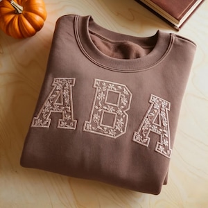 May include: A brown sweatshirt with the letters "ABA" embroidered on the front in a floral design. The sweatshirt is folded neatly on a wooden surface, with a small pumpkin and a book in the background.