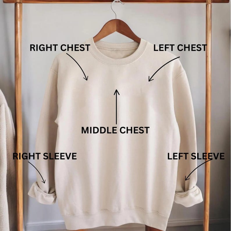 May include: A beige crew neck sweatshirt hangs on a wooden hanger. The image labels the sweatshirt's sections: Right Chest, Left Chest, Middle Chest, Right Sleeve, and Left Sleeve. The sweatshirt has a classic, casual design.
