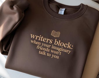 May include: A brown sweatshirt with the embroidered text "writers block: when your imaginary friends won't talk to you" and a book graphic. The sweatshirt has a crew neck and long sleeves.