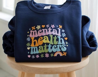Puede incluir: Sudadera azul marino con la frase "mental health matters" en letras coloridas de estilo retro. El diseño incluye flores, corazones y un arcoíris. La sudadera se muestra sobre un taburete de madera.