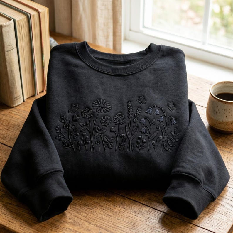 May include: A black sweatshirt with an embroidered floral design across the chest. The design includes various flowers, leaves, and butterflies. The sweatshirt is folded on a wooden surface, with a cup of coffee and books in the background.