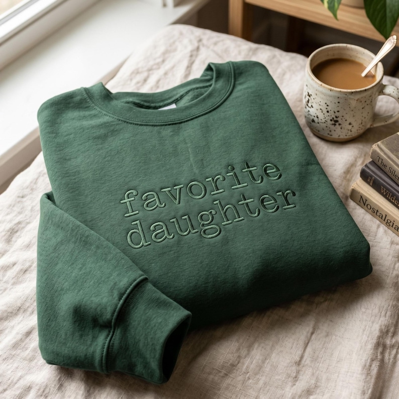 May include: A forest green sweatshirt with the words "favorite daughter" embroidered on the front. The sweatshirt is folded on a neutral-colored surface, with a coffee mug and books in the background.