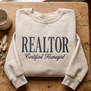 May include: A beige sweatshirt with the word "REALTOR" embroidered in dark gray, with "Certified Homegirl" below. The sweatshirt is folded on a wooden surface, with a ceramic mug and dried flowers in the background.