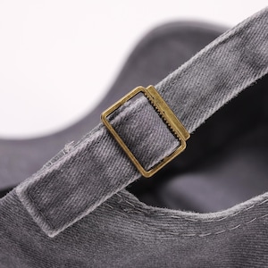 May include: Close-up of a gray baseball cap with a brass-colored metal buckle on the adjustable strap. The cap is made of a textured fabric, and the buckle is rectangular.