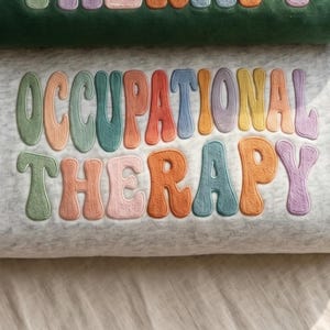 May include: A light gray heathered sweatshirt featuring the text OCCUPATIONAL THERAPY embroidered in a wavy, multi-colored font. The letters are stitched in shades of sage green, peach, teal, orange, blue, yellow, lavender, and pink.