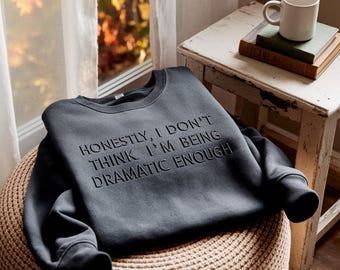 May include: Charcoal grey sweatshirt featuring the embossed phrase "HONESTLY, I DON'T THINK I'M BEING DRAMATIC ENOUGH". The garment is displayed on a woven pouffe, with a window and a small table holding a mug and books visible.