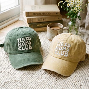 Puede incluir: Dos gorras de béisbol, una verde oliva y otra beige, se muestran sobre una superficie texturizada. Cada gorra presenta el texto bordado "TIRED MOMS CLUB" en blanco, con visera curva y correa ajustable. Parte de una colección de estilo de vida.