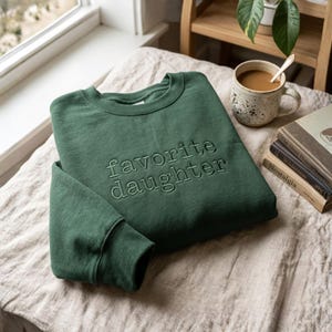 May include: A forest green sweatshirt with the phrase "favorite daughter" embroidered on the front. The sweatshirt is folded on a beige surface, with a cup of coffee and books in the background. The sweatshirt has a crew neck and long sleeves.