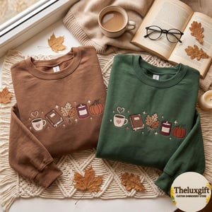 May include: Two crewneck sweatshirts, one brown and one dark green, each featuring an embroidered line of icons including a coffee mug with a heart, a closed book, a cluster of autumn leaves, a candle, and a pumpkin, accented with small stars.