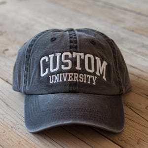 May include: A black, distressed, cotton baseball cap with white embroidered text that reads "CUSTOM UNIVERSITY".