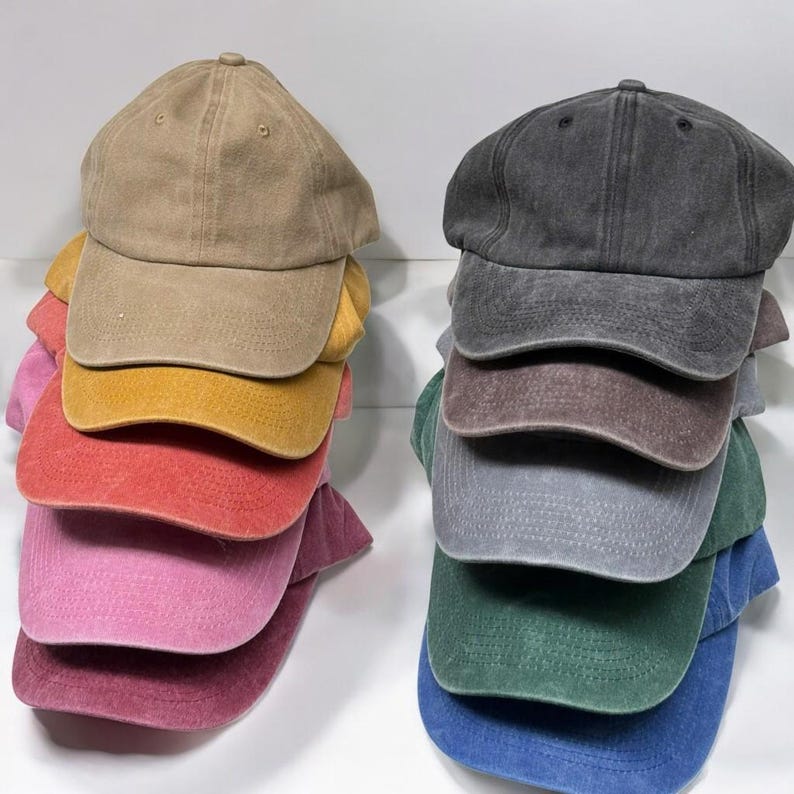 May include: Two stacks of baseball caps in various colors. The stack on the left includes tan, yellow, red, pink, and burgundy caps. The stack on the right features gray, brown, gray, green, and blue caps. Each cap has a curved brim and a top button.