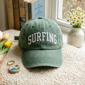 May include: A green baseball cap with the word "SURFING" embroidered in white across the front. The cap is made of a soft fabric and has a curved brim. A small keychain with a beach scene is visible in the foreground.