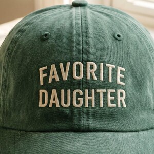 May include: A green baseball cap with the words "FAVORITE DAUGHTER" embroidered in white. The cap has a curved brim and a button on top. The hat is made of a soft, textured fabric.