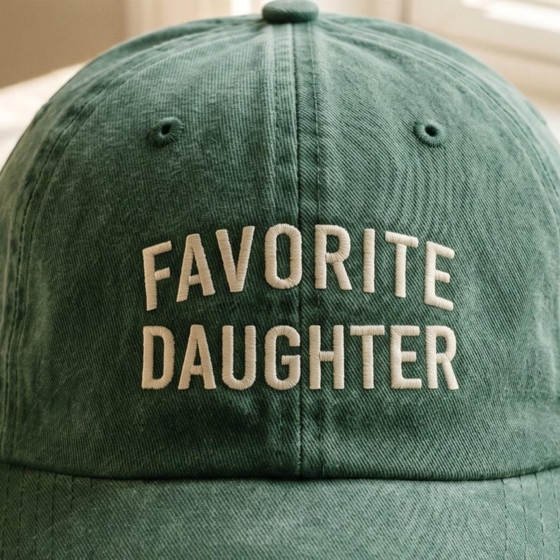 May include: A green baseball cap with the words "FAVORITE DAUGHTER" embroidered in white. The cap has a curved brim and a button on top. The hat is made of a soft, textured fabric.