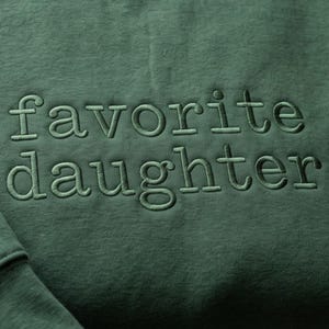 May include: Close-up of a forest green sweatshirt with the embroidered text "favorite daughter" in a matching green thread. The fabric has a textured appearance.