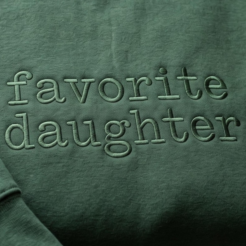 May include: Close-up of a forest green sweatshirt with the embroidered text "favorite daughter" in a matching green thread. The fabric has a textured appearance.