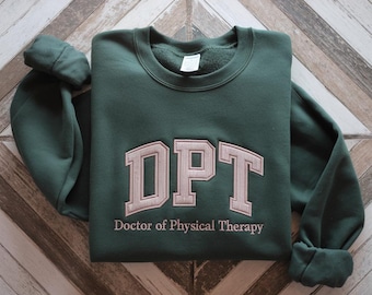 May include: A dark green sweatshirt with the embroidered letters "DPT" in a light tan colour. Below the letters is the text "Doctor of Physical Therapy". The sweatshirt is folded on a wooden surface.