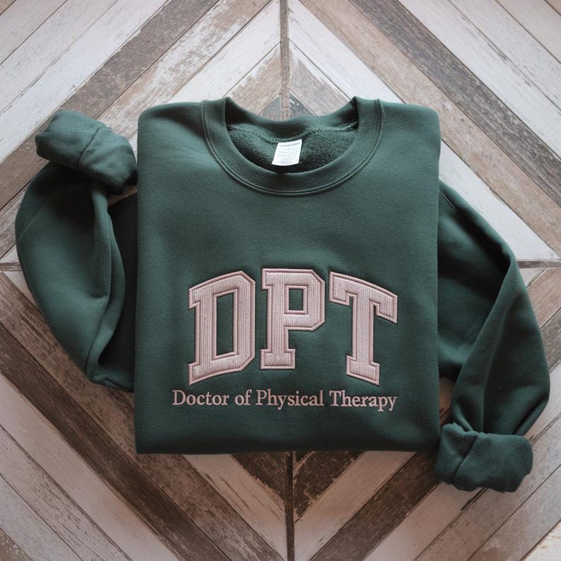 May include: A dark green sweatshirt with the embroidered letters "DPT" in a light tan colour. Below the letters is the text "Doctor of Physical Therapy". The sweatshirt is folded on a wooden surface.