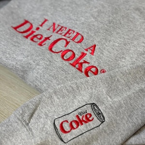 以下が含まれることがあります： 胸に「I NEED A Diet Coke」と赤い刺繍が施されたグレーのスウェットシャツ。袖には、赤い文字と黒いアウトラインでデザインされたダイエットコークの缶があります。