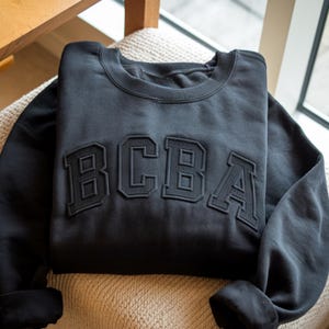May include: A black sweatshirt featuring the letters "BCBA" in a raised, textured design. The garment is folded on a beige, textured surface, with a window and wooden chair in the background.