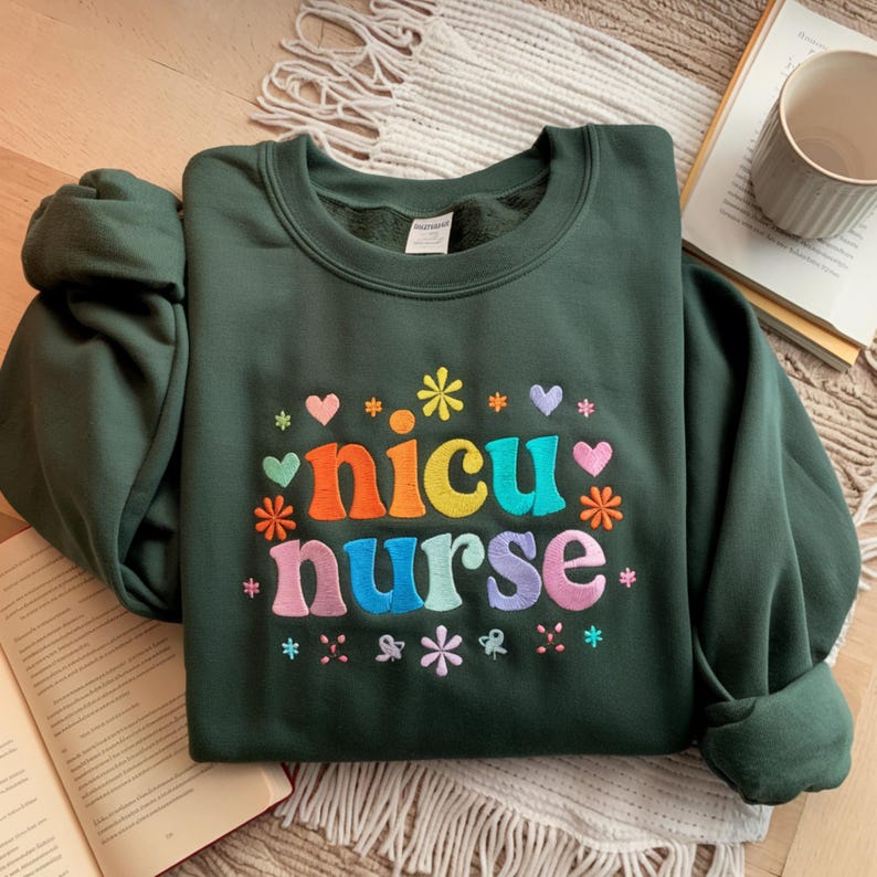 Puede incluir: Sudadera verde oscuro con el texto bordado "nicu nurse" en letras coloridas. El dise&ntilde;o incluye corazones y flores de varios colores. La sudadera est&aacute; doblada sobre una superficie estampada.