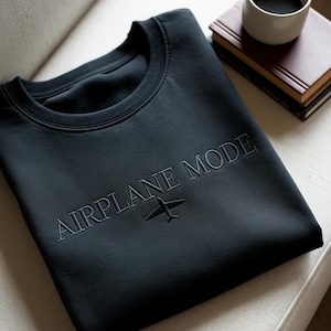 Puede incluir: Una sudadera negra de cuello redondo con la frase "AIRPLANE MODE" bordada en el pecho, junto con un peque&ntilde;o gr&aacute;fico de avi&oacute;n. La sudadera est&aacute; doblada sobre una silla blanca. Una taza de caf&eacute; y libros est&aacute;n en el fondo.