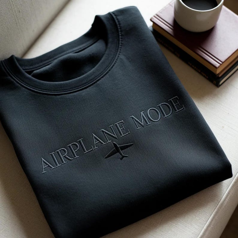 Puede incluir: Una sudadera negra de cuello redondo con la frase "AIRPLANE MODE" bordada en el pecho, junto con un peque&ntilde;o gr&aacute;fico de avi&oacute;n. La sudadera est&aacute; doblada sobre una silla blanca. Una taza de caf&eacute; y libros est&aacute;n en el fondo.