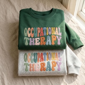 May include: A dark green sweatshirt and a light gray heather sweatshirt stacked together. Both feature the text OCCUPATIONAL THERAPY in a multi-colored, wavy, retro-style font with shades of pink, orange, yellow, blue, and purple letters.