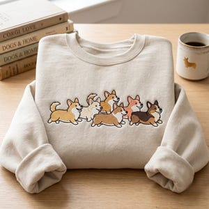 May include: A beige sweatshirt featuring an embroidered design of seven corgi dogs in various colors. The sweatshirt is displayed on a wooden surface, with books and a mug with a corgi silhouette visible in the background. The sweatshirt has a crew neck and long sleeves.