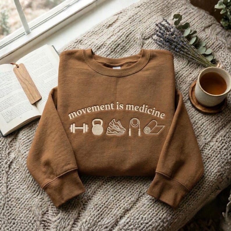 May include: A brown sweatshirt with the phrase "movement is medicine" embroidered in white, along with fitness-related icons. The sweatshirt is displayed on a textured, neutral-colored blanket, with a book and a cup of tea nearby.