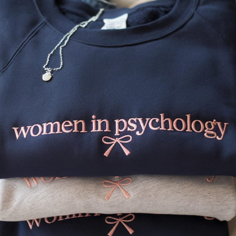 May include: A stack of navy blue and gray sweatshirts embroidered with the words "women in psychology" in a pink script font. Each sweatshirt has a pink bow graphic below the text. A silver necklace with a small pendant rests on the top sweatshirt.