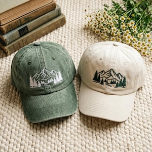 May include: Two baseball caps with mountain and tree embroidery. One cap is olive green, the other is beige. The caps are made of a soft fabric and have a curved brim. The caps are a casual accessory.