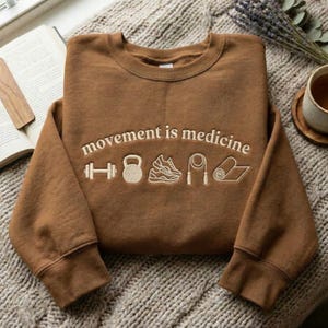 May include: A brown sweatshirt with the embroidered text "movement is medicine" and fitness-related icons. The icons include a dumbbell, kettlebell, running shoe, jump rope, and yoga mat. The sweatshirt is displayed on a textured, neutral-colored surface.