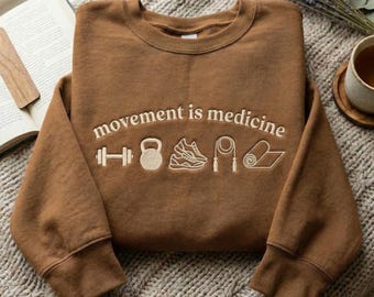 May include: A brown sweatshirt featuring the embroidered phrase "movement is medicine" and fitness icons. The icons include a dumbbell, kettlebell, running shoe, skipping rope, and yoga mat. The sweatshirt is presented on a textured, neutral-toned surface.