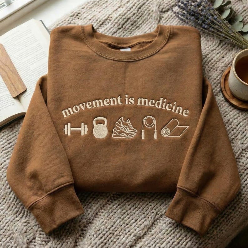 May include: A brown sweatshirt with the embroidered text "movement is medicine" and fitness-related icons. The icons include a dumbbell, kettlebell, running shoe, jump rope, and yoga mat. The sweatshirt is displayed on a textured, neutral-colored surface.