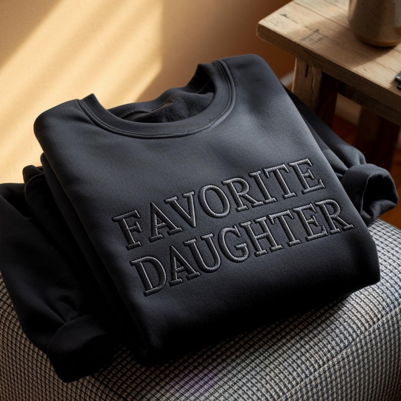 May include: A black sweatshirt with the embroidered text "FAVORITE DAUGHTER" in a matching dark gray color. The sweatshirt is folded and resting on a patterned surface, with a wooden table and a mug in the background.