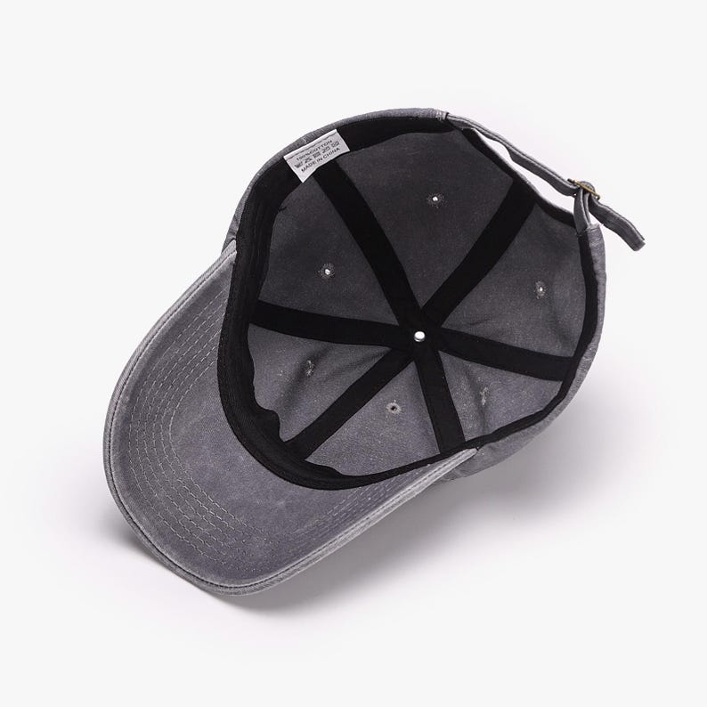 May include: A gray baseball cap with a curved brim, viewed from the inside. The cap features a six-panel design with a black interior lining and a fabric strap for adjustment. The cap has a faded, worn appearance.