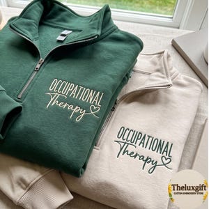 May include: A dark green quarter-zip sweatshirt and a cream quarter-zip sweatshirt, both featuring embroidered text that reads Occupational Therapy with a small heart icon. The green sweatshirt has cream embroidery, while the cream sweatshirt has green embroidery.