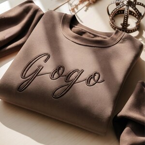 May include: A folded brown sweatshirt with the embroidered word "Gogo" in a cursive font. The sweatshirt is a solid color with a crew neck. Additional brown fabric is visible in the background, along with a decorative crown.